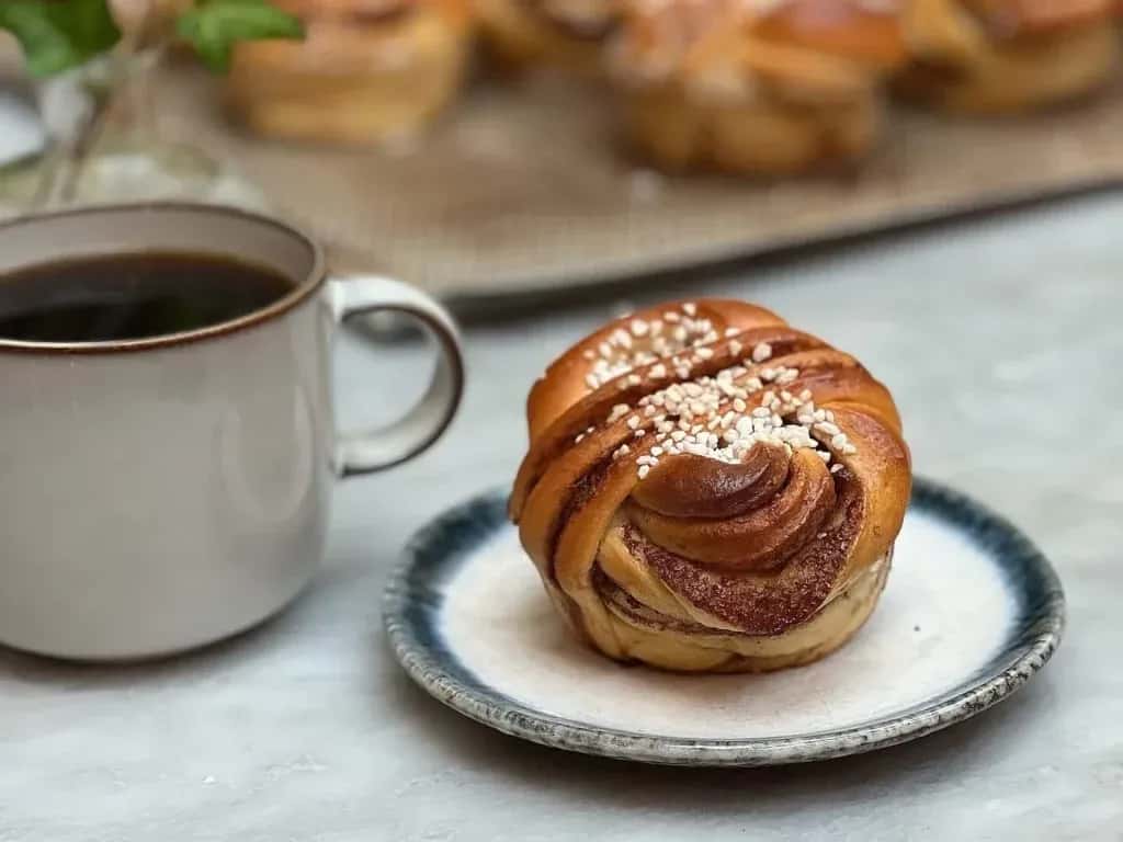 Kanelbulle med pärlsocker på fat, kaffe i mugg. Fler bullar syns i bakgrunden