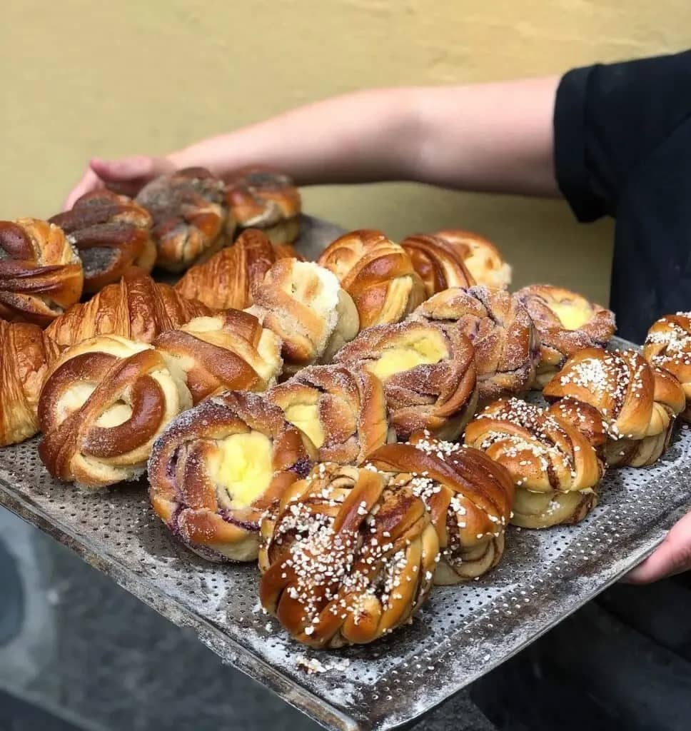 En person håller en plåt med nybakade kanelbullar, croissanter och vaniljkrämsbullar, många med pärlsocker