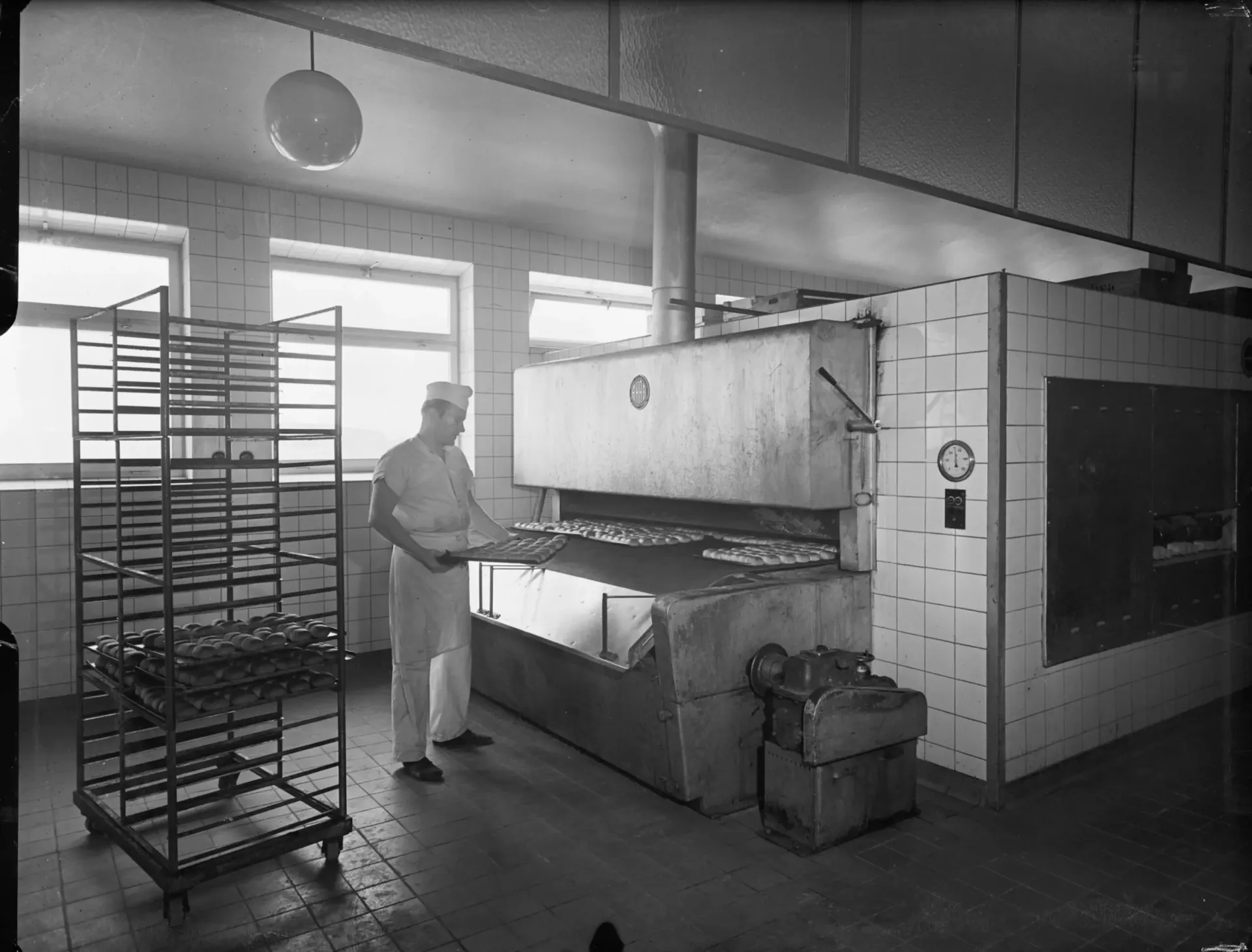 Bagare placerar plåtar med runda bröd i en stor industriugn i ett kaklat bageri. En vagn med bröd står bredvid